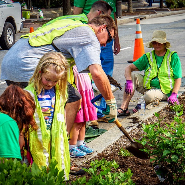 Photo of a neighborhood cleanup event. People of all ages are planting new plants on a sidewalk strip. Some are wearing bright yellow safety vests. 