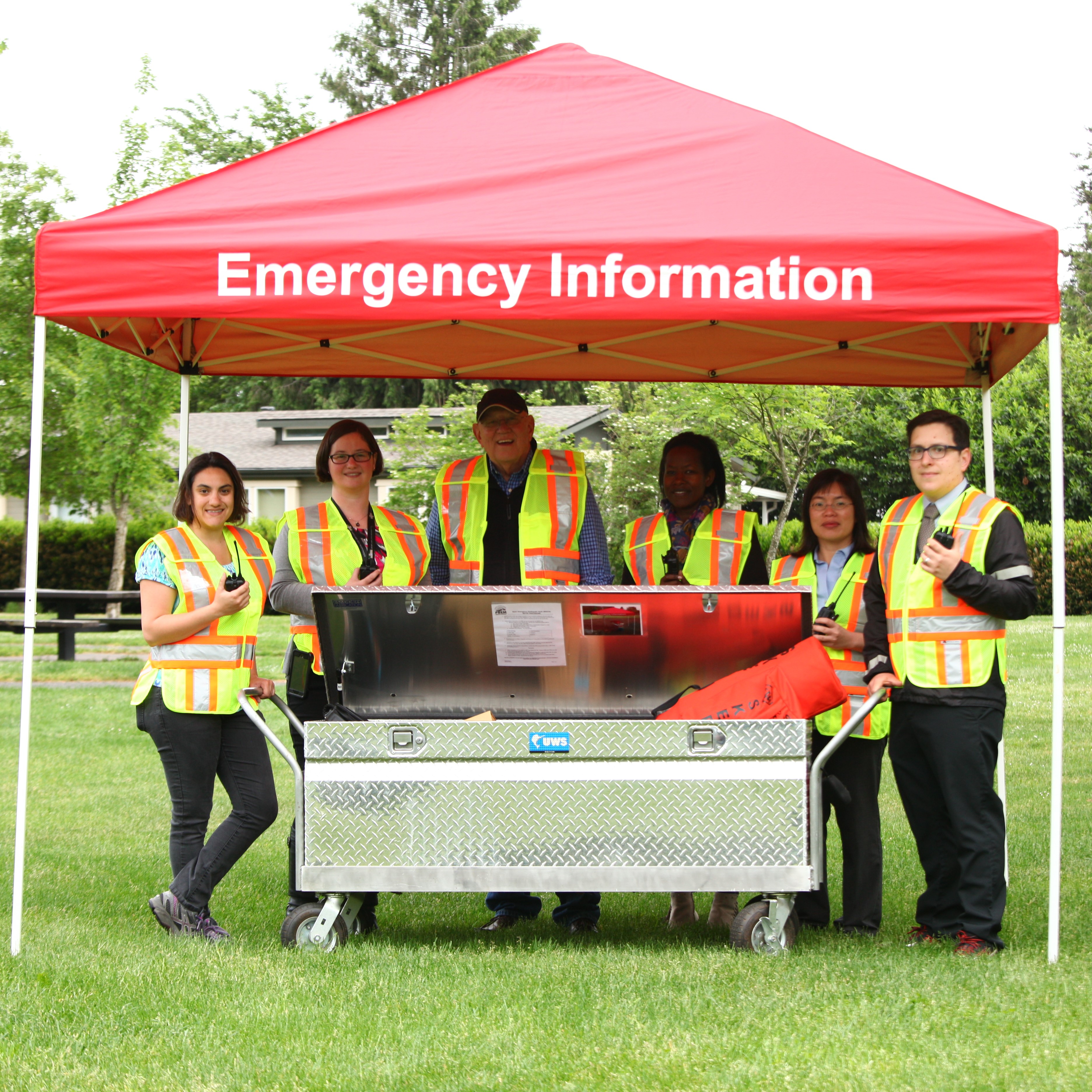 Volunteers in yellow and orange safety vests stand under a red tent labeled "Emergency Information." They are holding radios and standing behind a shiny silver box with the lid partially ajar. 
