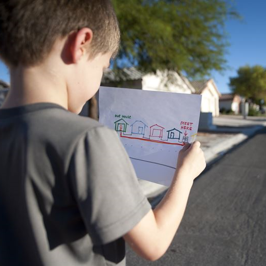 Young Caucasian boy stands on a suburban street. He is holding a hand-drawn map of his neighborhood with his house and a meeting spot labeled. 