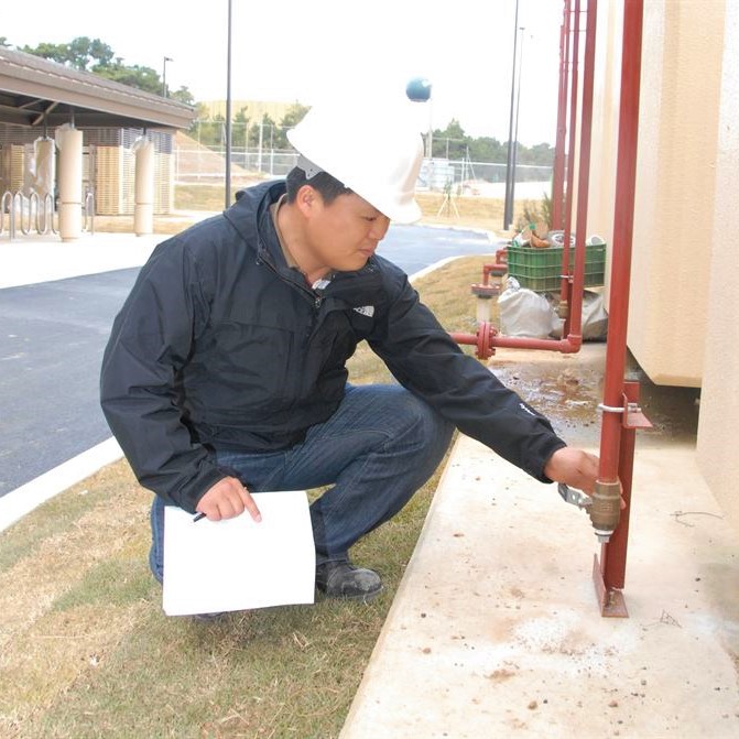Building inspector wearing hard hat and carrying paper and pen is kneeling on one knee and looking at a pipe.