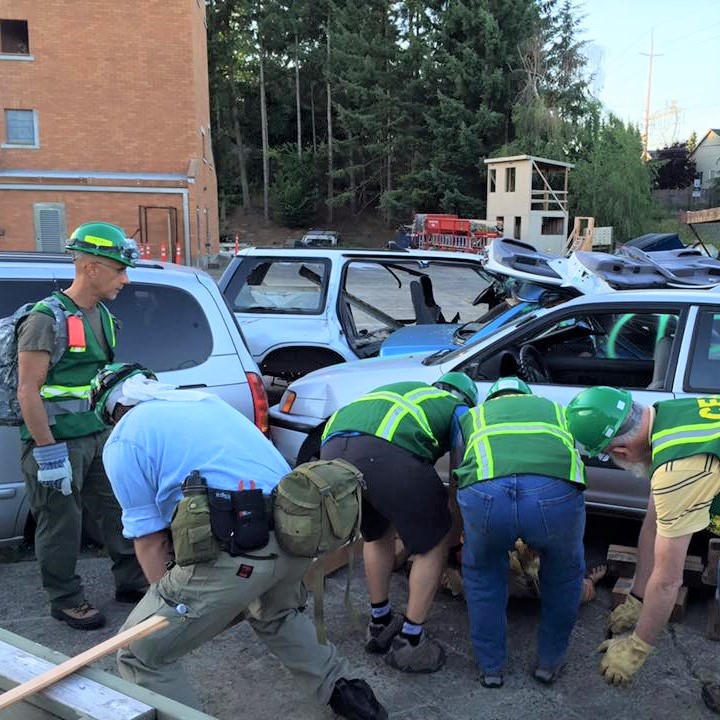 Clark County CERT volunteers get trained on cribbing. Four people are attempting to rescue a mannequin by lifting a car with wood blocks. A 5th person who is the trainer observes. 