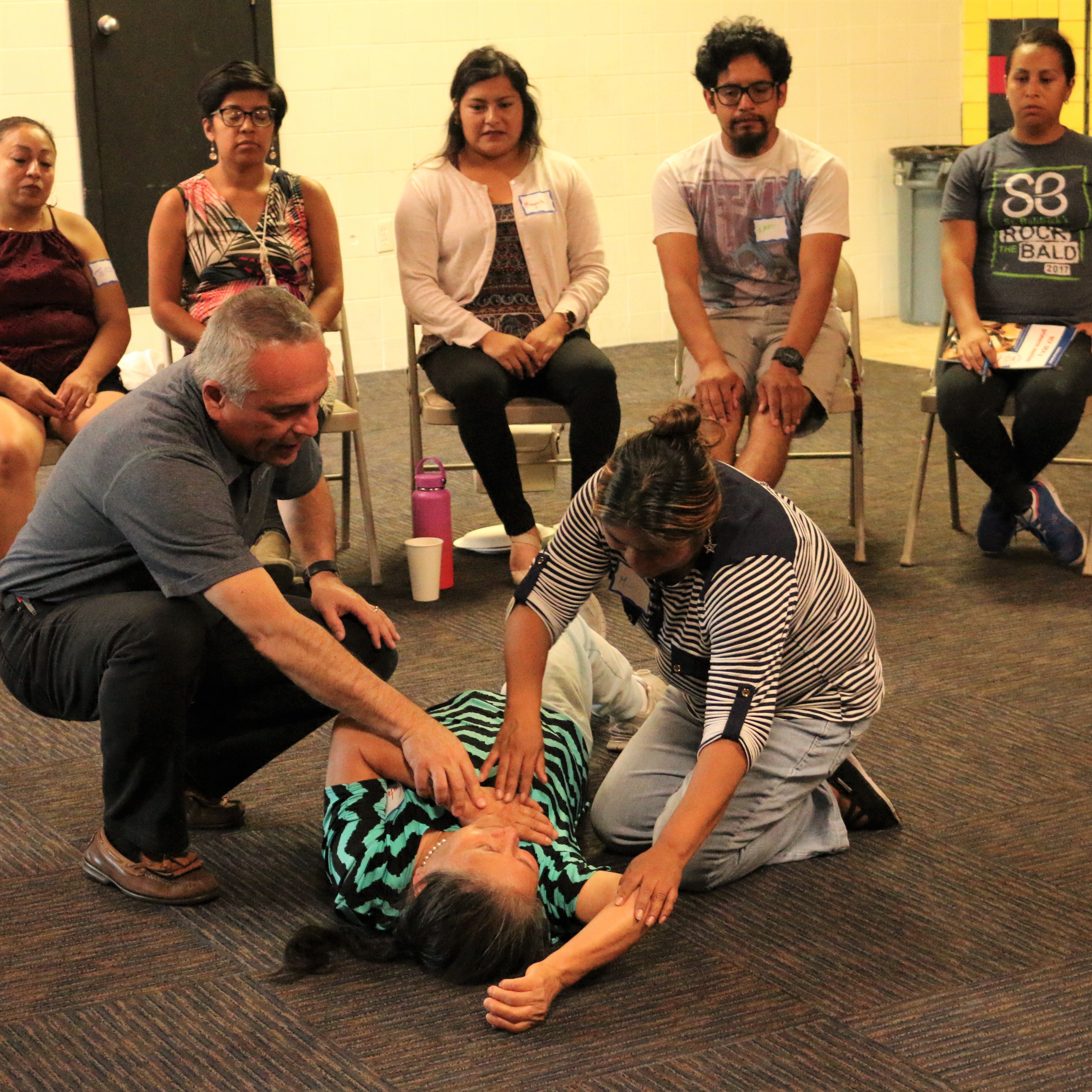 LISTOS volunteers receive CPR training in Portland's Cully neighborhood. An instructor shows a participant how to check the airway of victim volunteer. Several participants sit behind them and watch.