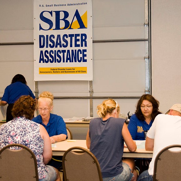 Two women in blue shirts sit across the table from people who are applying for post-disaster assistance. The sign behind them says "Small Business Association Disaster Assistance." 